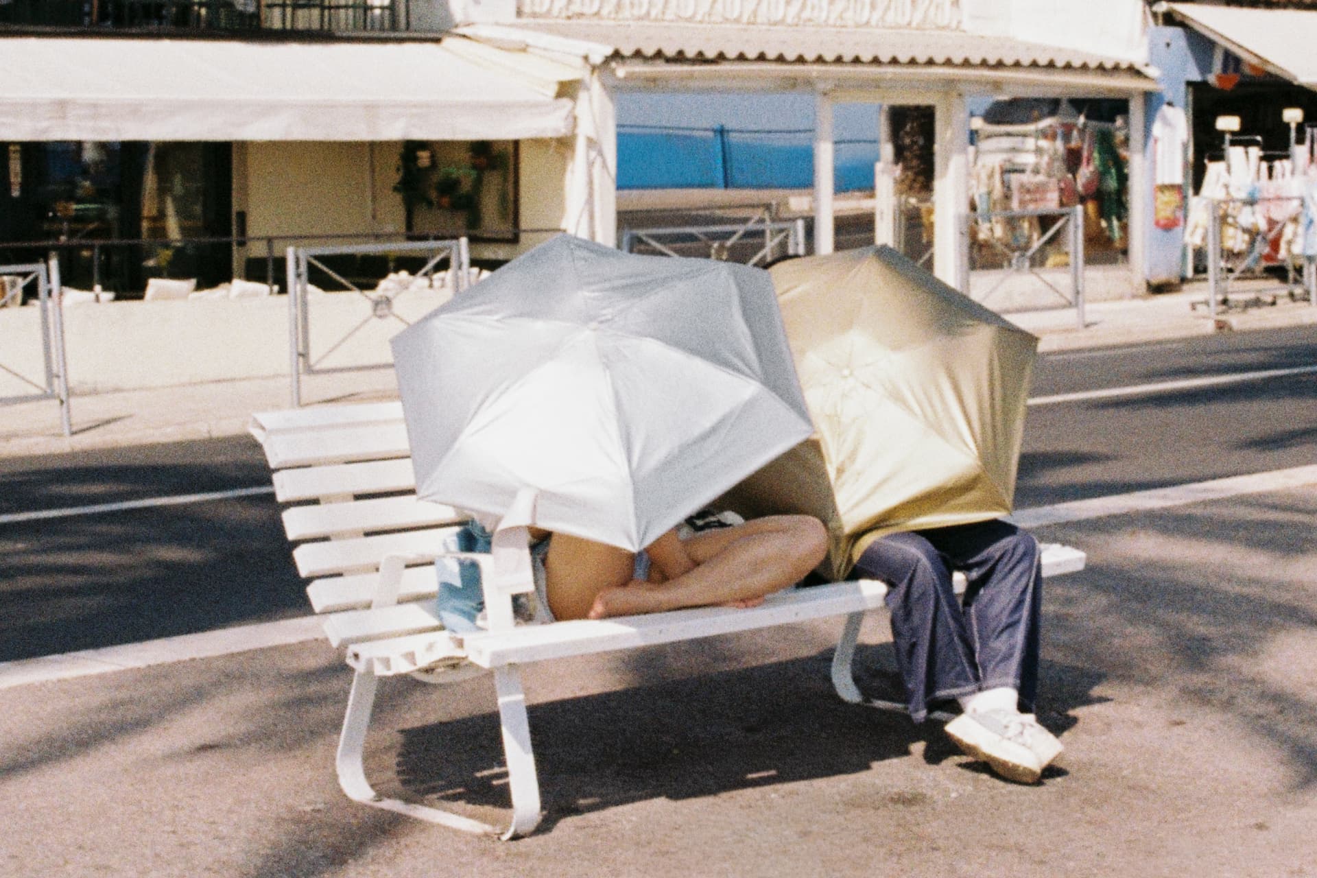 Two people hiding under umbrellas on a white bench by the sea