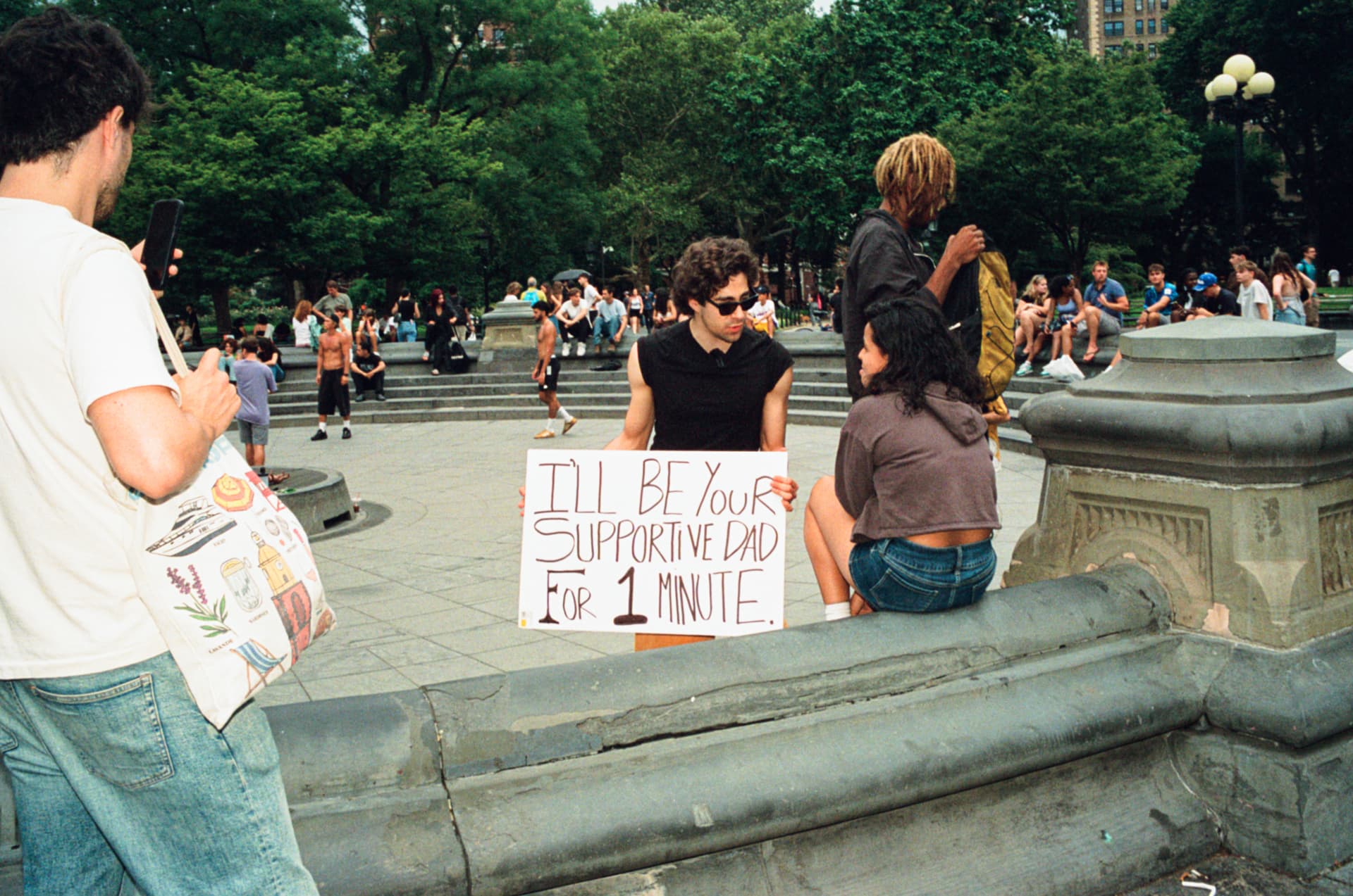 Man holding a sign reading I'll be your supportive dad for 1 minute in a park