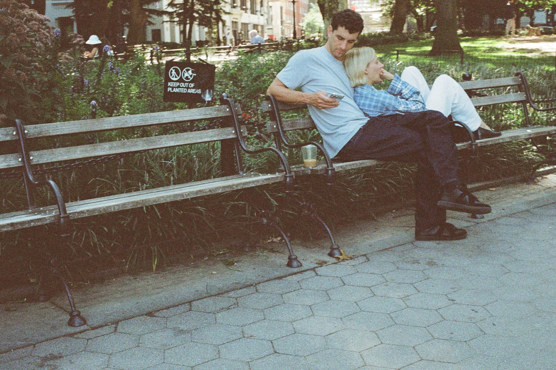 Couple leaning together on a wooden park bench beside a keep out of planted areas sign