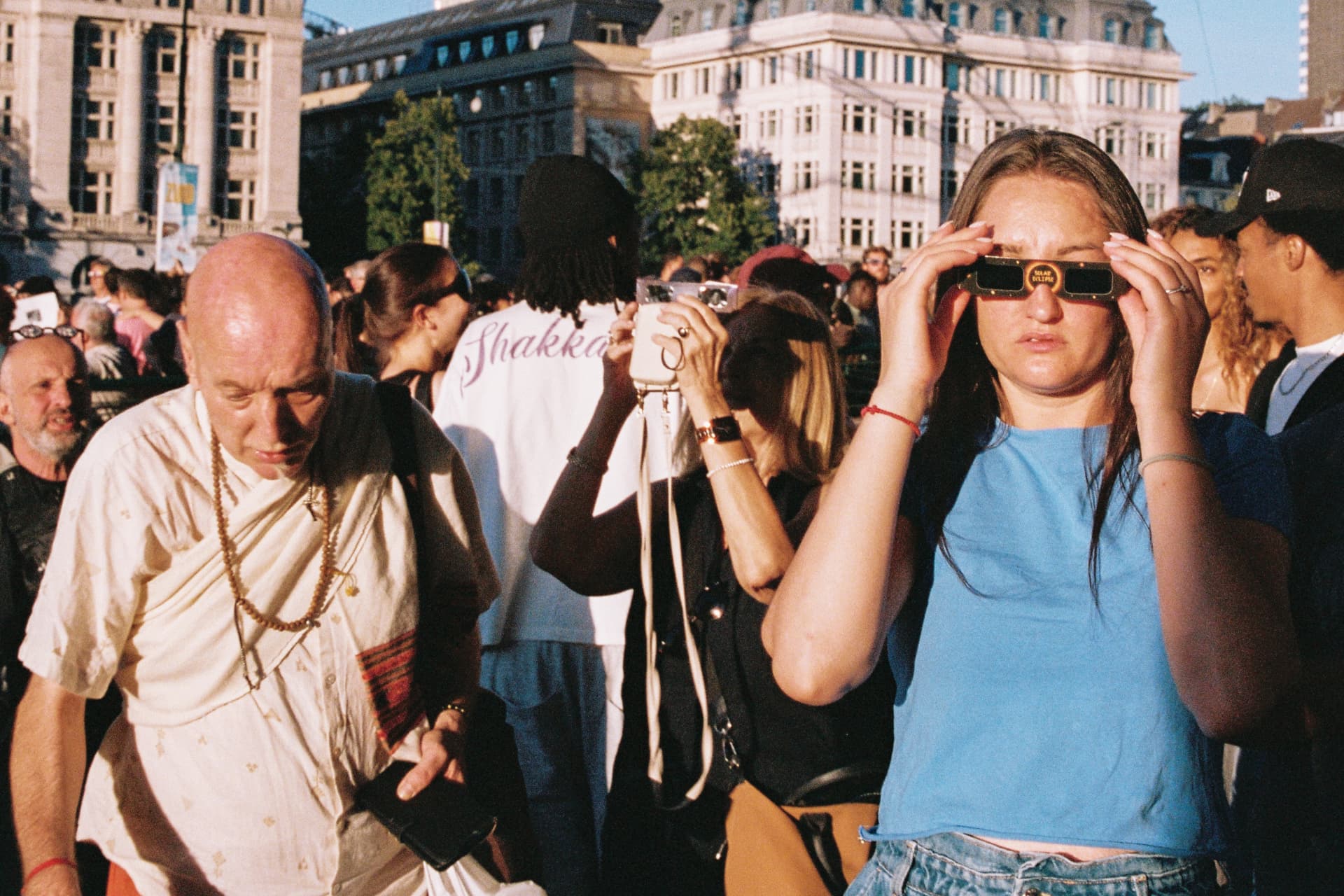 Woman in a blue t-shirt looking through solar eclipse glasses in a crowd, with a monk in white walking past