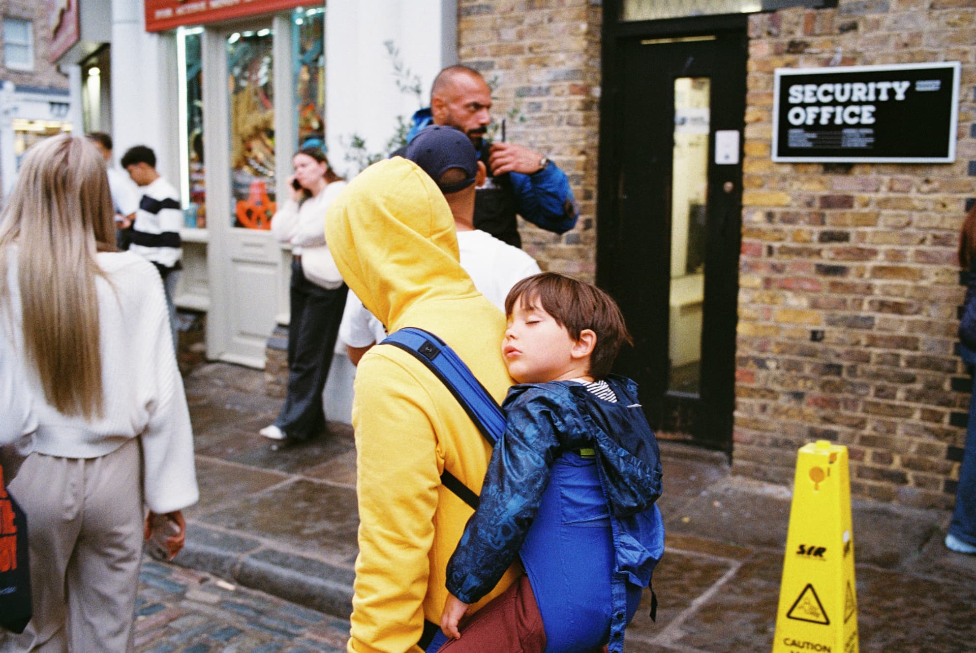 Toddler asleep in a blue child carrier on a man's back in a yellow hoodie, near a Security Office sign