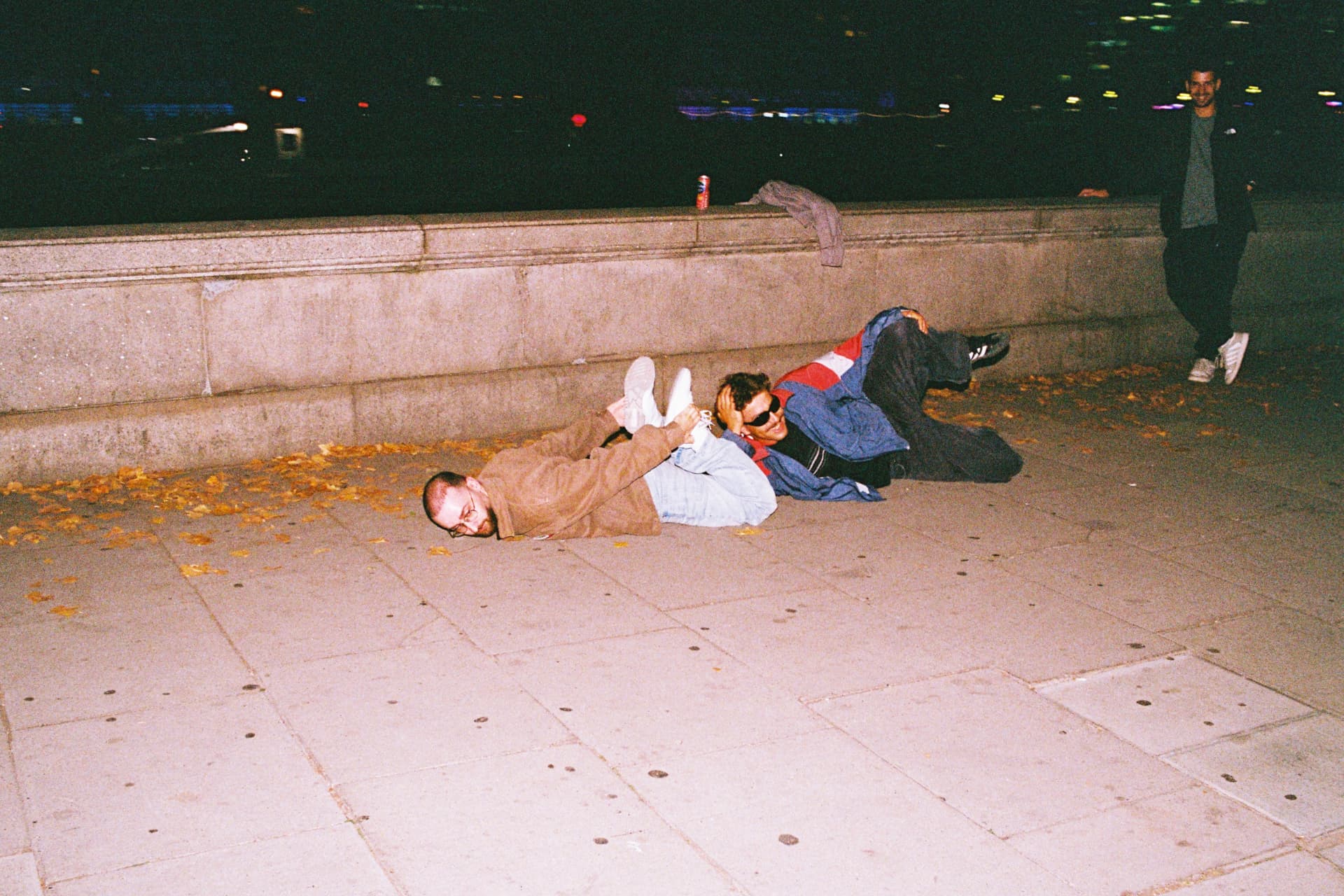 Two friends lying on the pavement laughing by the river at night, autumn leaves around them