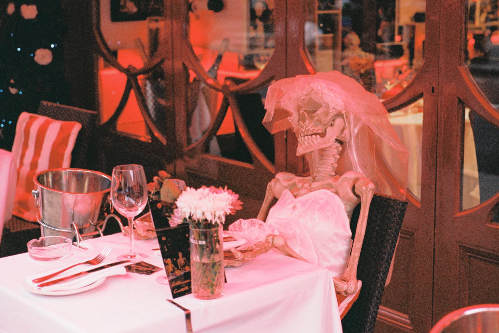 Skeleton bride in a veil and wedding dress seated at a red-lit restaurant table with wine glasses and white flowers