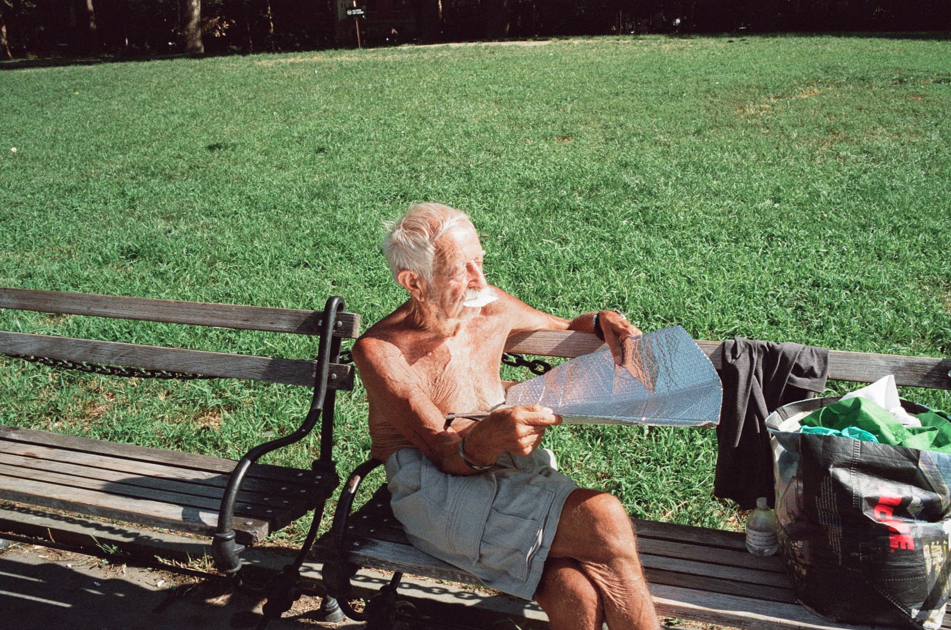 Shirtless older man reading a newspaper on a park bench