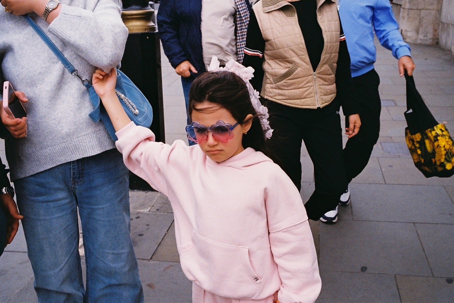 Girl in a pink hoodie and flower-shaped sunglasses holding a blue shoulder bag up on a busy street