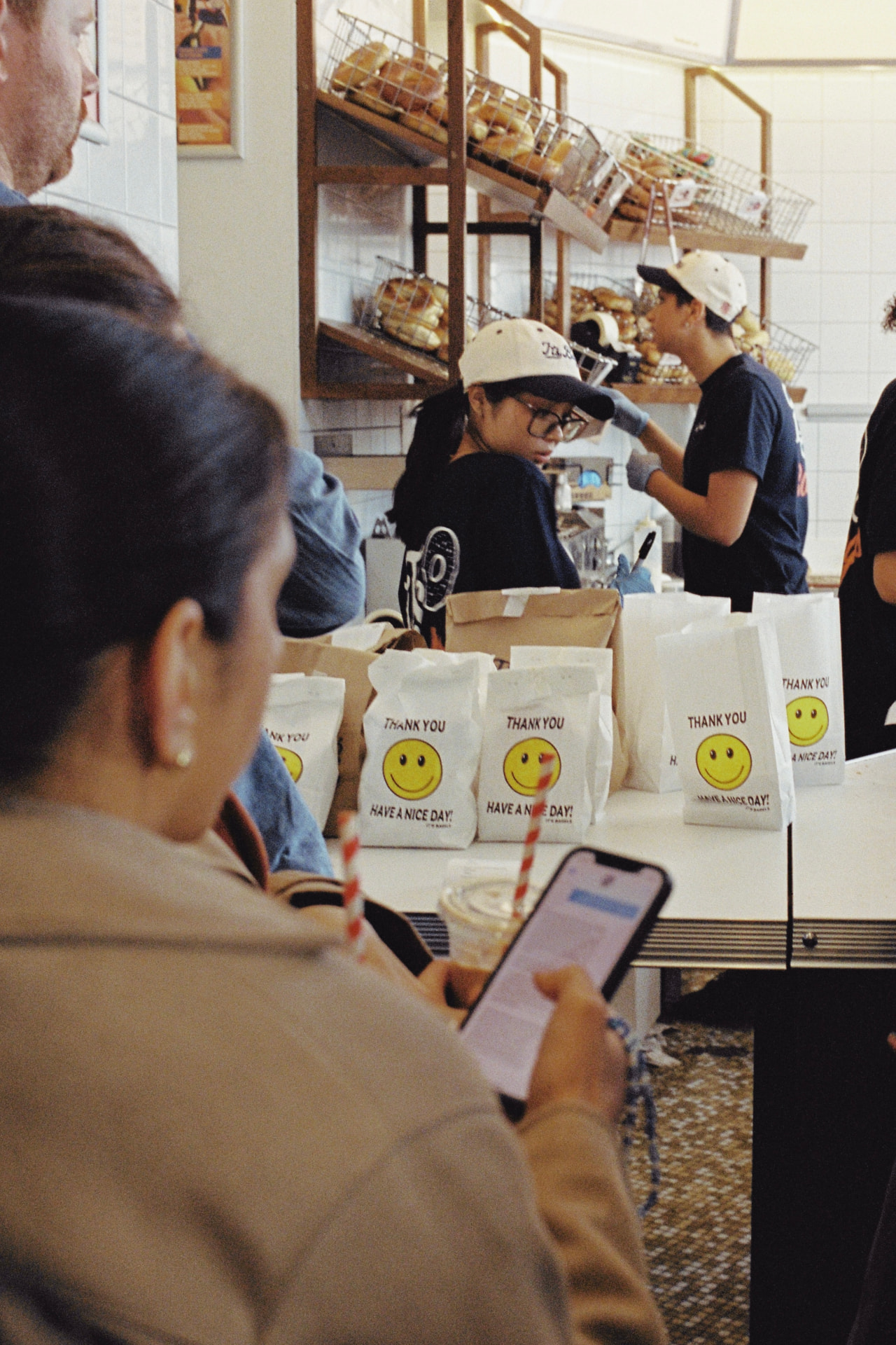 Staff serving customers behind a bakery counter lined with white paper bags printed with smiley faces and thank you, have a nice day
